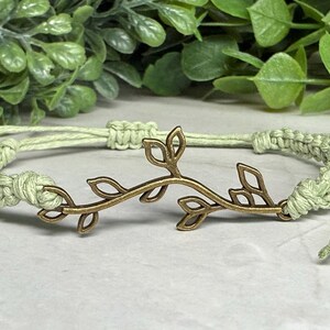May include: A light green macrame bracelet with a bronze-colored leaf design. The bracelet is made of knotted cord and features an adjustable closure. The leaf design is centered on the bracelet.