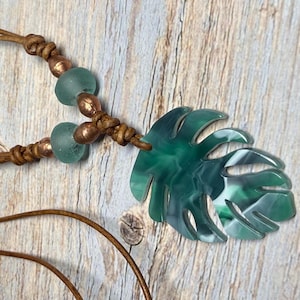 May include: A handcrafted necklace featuring a green and white leaf pendant, sea glass beads, and copper-colored beads. The pendant is a stylized monstera leaf. The necklace is strung on a brown leather cord.