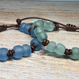 May include: Two handmade bracelets with sea glass beads in shades of blue and green. The beads are strung on brown leather cords with adjustable knots. The bracelets are displayed on a wooden surface.
