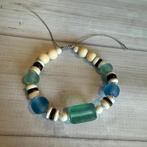 May include: A handmade bracelet featuring a mix of natural wood beads, sea glass, and dark brown and white accent beads. The bracelet includes round and rectangular sea glass pieces in shades of blue and green, strung on a gray cord.