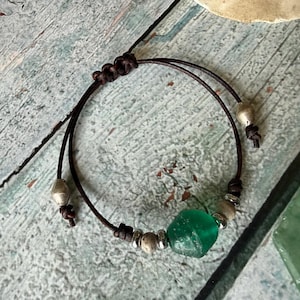 May include: A dark brown leather cord bracelet featuring a single translucent green sea glass bead, accented with small metallic silver-toned beads and textured stone-like spacers, secured with an adjustable sliding knot closure mechanism.