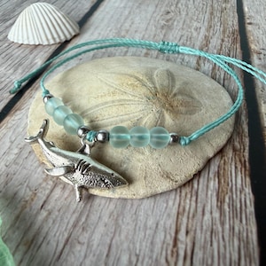 May include: A light blue cord bracelet featuring a silver-toned shark charm, accented with six translucent seafoam green frosted beads and small silver-toned metal spacers, finished with an adjustable sliding knot closure mechanism.