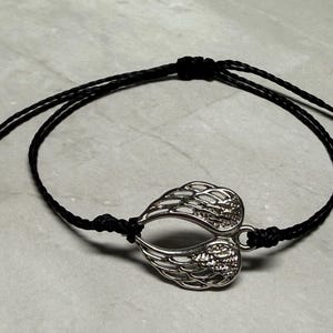 May include: A silver-coloured wing charm bracelet with a black cord. The charm is a detailed depiction of two wings joined together. The bracelet is knotted for adjustability and is displayed on a light grey surface.