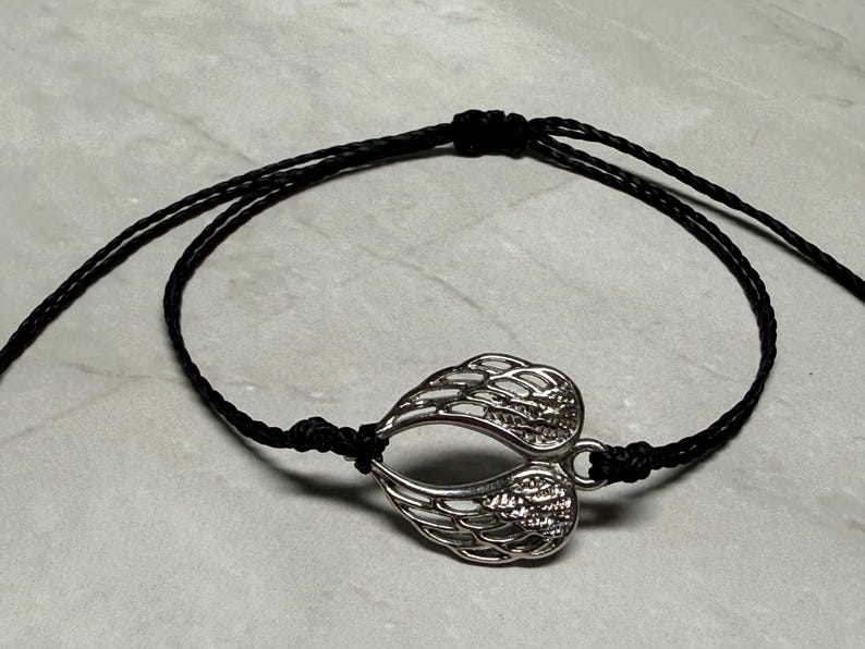 May include: A silver-coloured wing charm bracelet with a black cord. The charm is a detailed depiction of two wings joined together. The bracelet is knotted for adjustability and is displayed on a light grey surface.