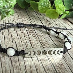 May include: A handmade bracelet featuring white moonstone beads and a silver-colored bar with crescent moon cutouts. The bracelet is strung on a black cord, with adjustable sliding knots for a comfortable fit. The bracelet is displayed on a wooden surface.