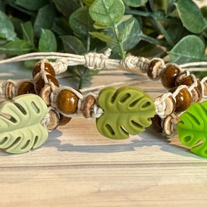 May include: Three macrame bracelets with wooden beads and a leaf-shaped charm. The bracelets are made with a natural-colored cord and feature brown wooden beads and a green leaf charm. The bracelets are displayed on a light-colored wooden surface.