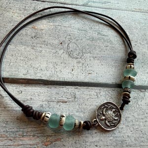 May include: A dark brown leather cord necklace featuring a circular silver-toned pendant with a sea turtle relief design. The necklace is accented with light green sea glass beads and small, textured, off-white cylindrical spacer beads.
