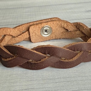 May include: Braided brown leather bracelet with a snap closure. The bracelet features a woven design with a light brown interior and a darker brown exterior. The bracelet is approximately 0.75 inches wide.