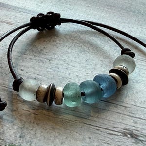 May include: A bracelet featuring a dark brown leather cord with an adjustable sliding knot closure. The cord is strung with a series of smooth, frosted sea glass beads in shades of light blue, teal, and clear, interspersed with small, round, dark brown and cream-colored wooden spacer beads.