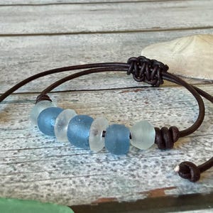 May include: A bracelet featuring a dark brown leather cord with an adjustable sliding knot closure. The center is adorned with seven smooth, frosted sea glass beads in alternating shades of light blue, medium blue, and translucent white.