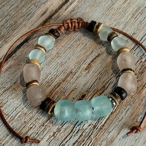 K&ouml;nnte beinhalten: Ein handgefertigtes Armband mit Seeglasperlen in Hellblau und Rosa, gemischt mit Holz- und dunkelbraunen Perlen. Das Armband ist an einer braunen Lederschnur aufgereiht, die an den Enden geknotet ist.