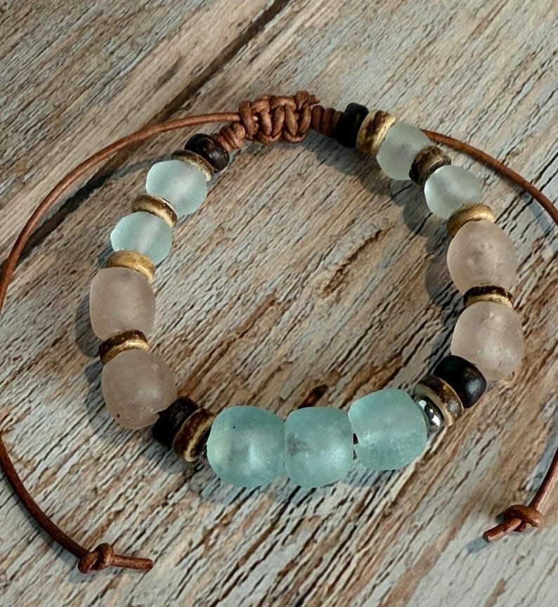K&ouml;nnte beinhalten: Ein handgefertigtes Armband mit Seeglasperlen in Hellblau und Rosa, gemischt mit Holz- und dunkelbraunen Perlen. Das Armband ist an einer braunen Lederschnur aufgereiht, die an den Enden geknotet ist.