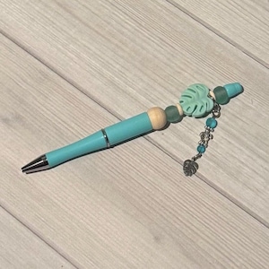 May include: A turquoise pen with silver accents and a decorative end. The end features a light green leaf-shaped charm, beads, and a small silver leaf charm. The pen is a writing accessory.