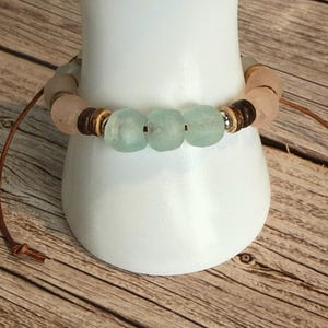 K&ouml;nnte beinhalten: Ein handgefertigtes Armband mit hellblauen und rosa Seeglasperlen, kombiniert mit Holz- und Silberelementen. Das Armband ist an einer braunen Lederschnur befestigt und pr&auml;sentiert ein nat&uuml;rliches, erdiges Design.