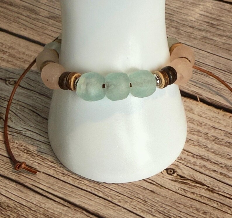 K&ouml;nnte beinhalten: Ein handgefertigtes Armband mit hellblauen und rosa Seeglasperlen, kombiniert mit Holz- und Silberelementen. Das Armband ist an einer braunen Lederschnur befestigt und pr&auml;sentiert ein nat&uuml;rliches, erdiges Design.