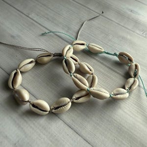 May include: Two cowrie shell bracelets. Each bracelet is made of cream-colored shells strung together with a thin, light-colored cord. One bracelet is connected with a gray cord, and the other with a teal cord. The bracelets are displayed on a light wood surface.