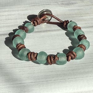 May include: A handmade bracelet featuring sea glass beads in a soft teal color, interspersed with brown leather knots. The bracelet is secured with a silver-toned button clasp. The design evokes a beachy, bohemian aesthetic.
