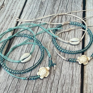 May include: Several handmade bracelets with a beach theme. The bracelets are made with blue and white beads, seashells, and turtle charms. The bracelets are on a wooden surface.