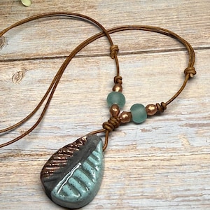 May include: A handmade necklace featuring a ceramic leaf pendant with a brown and teal design. The pendant is suspended from a brown leather cord, accented with teal beads and copper-colored beads. The necklace is displayed on a wooden surface.