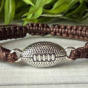 May include: A brown macrame bracelet featuring a silver football charm. The bracelet is made of woven brown cord and has an adjustable closure. The football charm is detailed with textured lines and a classic design.