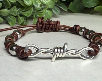 Könnte beinhalten: Braunes Lederarmband mit silbernen Perlen und einem silbernen Stacheldraht-Design in der Mitte. Das Armband ist verstellbar und hat einen geknoteten Verschluss. Die Perlen sind gleichmäßig verteilt.
