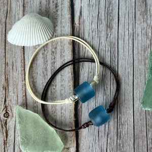 May include: Two adjustable bracelets with blue glass beads. One bracelet has a cream-colored cord, and the other has a dark brown cord. A white seashell and two pieces of sea glass are also visible on a wooden surface.