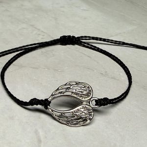 May include: A silver-coloured wing charm bracelet with a black cord. The charm is shaped like two wings joined together. The bracelet is adjustable with sliding knots.