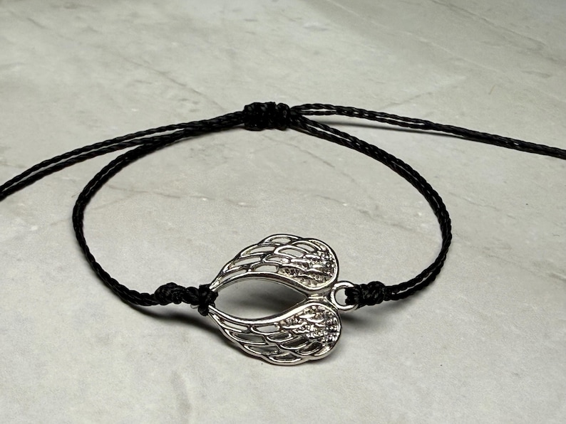 May include: A silver-coloured wing charm bracelet with a black cord. The charm is shaped like two wings joined together. The bracelet is adjustable with sliding knots.