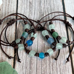 May include: A collection of three bracelets made from dark brown leather cords and frosted sea glass beads in shades of light green, dark green, and blue. Each bracelet features a series of beads separated by knots along the leather cord.