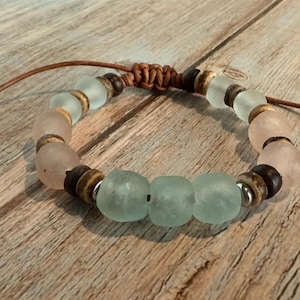 K&ouml;nnte beinhalten: Ein handgefertigtes Armband mit mattierten Glasperlen in Rosa und Meeresgr&uuml;n, kombiniert mit dunklen Holzperlen und silberfarbenen Akzenten. Das Armband ist an einem braunen Lederband befestigt.