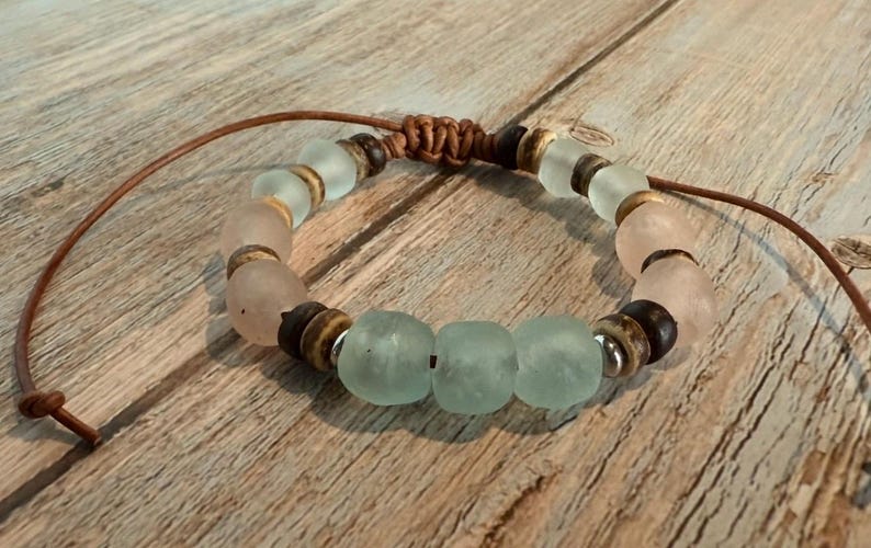 K&ouml;nnte beinhalten: Ein handgefertigtes Armband mit mattierten Glasperlen in Rosa und Meeresgr&uuml;n, kombiniert mit dunklen Holzperlen und silberfarbenen Akzenten. Das Armband ist an einem braunen Lederband befestigt.