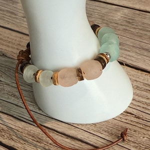 K&ouml;nnte beinhalten: Ein handgefertigtes Armband mit rosa und mintgr&uuml;nen Seeglasperlen, Holzperlen und braunen Perlen. Es ist auf einer braunen Lederschnur aufgef&auml;delt, die am Ende verknotet ist. Das Armband wird auf einer wei&szlig;en Form pr&auml;sentiert.
