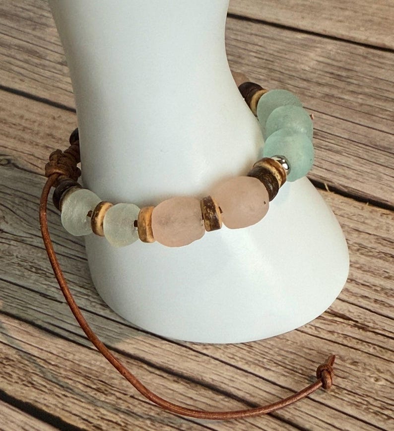 K&ouml;nnte beinhalten: Ein handgefertigtes Armband mit rosa und mintgr&uuml;nen Seeglasperlen, Holzperlen und braunen Perlen. Es ist auf einer braunen Lederschnur aufgef&auml;delt, die am Ende verknotet ist. Das Armband wird auf einer wei&szlig;en Form pr&auml;sentiert.