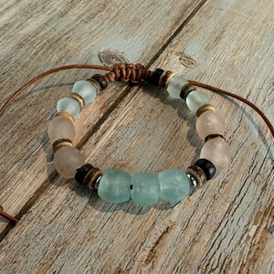 K&ouml;nnte beinhalten: Ein handgefertigtes Armband mit Seeglasperlen in Hellblau und Rosa, kombiniert mit Holz- und Metallelementen. Es ist auf einer braunen Lederschnur aufgef&auml;delt, mit verstellbaren Knoten. Ca. 18 cm lang.