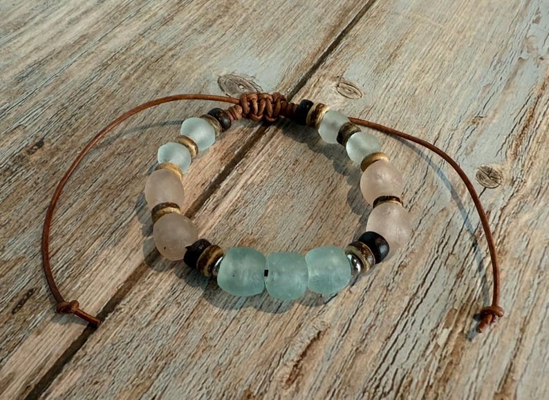 K&ouml;nnte beinhalten: Ein handgefertigtes Armband mit Seeglasperlen in Hellblau und Rosa, kombiniert mit Holz- und Metallelementen. Es ist auf einer braunen Lederschnur aufgef&auml;delt, mit verstellbaren Knoten. Ca. 18 cm lang.
