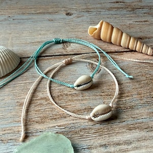 May include: Two handmade seashell bracelets with adjustable cords in light blue and cream. A white seashell and a spiral seashell are also visible. The bracelets feature a cowrie shell as a focal point, perfect for beach-themed jewelry.