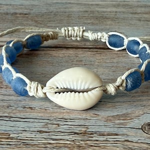 May include: A handmade bracelet featuring blue glass beads and a white seashell centerpiece. The beads are strung on a natural-colored cord, creating a nautical-themed accessory.