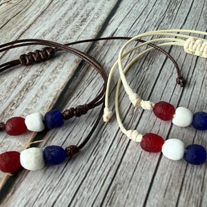 May include: Two handmade bracelets with red, white, and blue beads. One bracelet has a brown cord, and the other has a cream-colored cord. The beads are strung on the cord and secured with knots. The bracelets are displayed on a wooden surface.