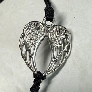 May include: Silver-coloured pendant shaped like angel wings, hanging from a black cord. The wings have detailed feathering and are joined at the base. The cord is knotted above and below the pendant.