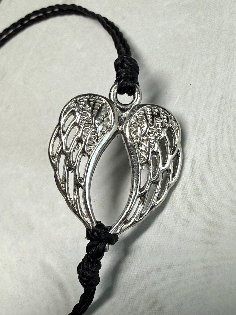 May include: Silver-coloured pendant shaped like angel wings, hanging from a black cord. The wings have detailed feathering and are joined at the base. The cord is knotted above and below the pendant.