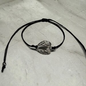 May include: A black cord bracelet with a silver-coloured charm shaped like angel wings. The bracelet has adjustable sliding knots and small silver beads at the ends. The wings are detailed with feather-like patterns.