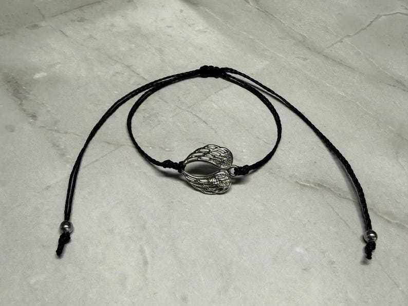 May include: A black cord bracelet with a silver-coloured charm shaped like angel wings. The bracelet has adjustable sliding knots and small silver beads at the ends. The wings are detailed with feather-like patterns.