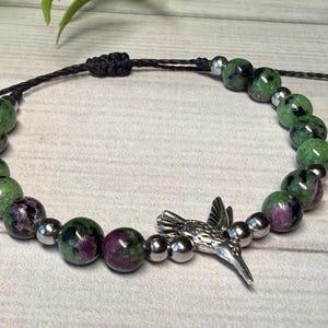 May include: Adjustable bracelet featuring green and purple gemstone beads and silver-colored accent beads. A silver hummingbird charm is centered on the bracelet. The bracelet is strung on a black cord.