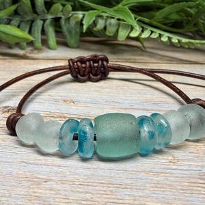 May include: A handmade bracelet featuring sea glass beads in shades of blue and clear, strung on a brown leather cord. The bracelet has a sliding knot closure for adjustable sizing. The sea glass beads are smooth and rounded.