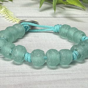 May include: A sea glass bracelet featuring round, frosted, light blue beads. The beads are strung on a matching light blue cord, with a silver clasp. The bracelet is a handmade jewelry item.