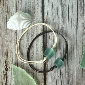May include: Two bracelets featuring smooth, frosted sea glass beads in a soft teal color. One bracelet is crafted with a light cream-colored cord, while the other is made with a dark brown cord. Both bracelets include adjustable knots for sizing.