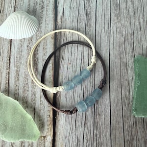 May include: Two bracelets with light blue beads. One bracelet is brown, the other is cream-colored. The bracelets are on a wooden surface with a seashell and sea glass.