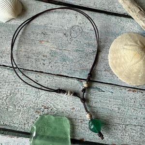 May include: A dark brown leather cord necklace featuring a single green glass bead pendant at the center. The necklace is accented with small metallic silver-toned beads and knots along the cord, creating a simple, adjustable lariat-style design.