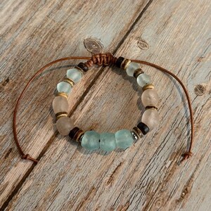 K&ouml;nnte beinhalten: Ein handgefertigtes Armband mit einer Mischung aus hellblauen, klaren und braunen Perlen. Das Armband ist an einer braunen Kordel mit einem Schiebeknotenverschluss befestigt. Die Perlen haben verschiedene Formen und Gr&ouml;&szlig;en und schaffen einen einzigartigen, b&ouml;hmischen Stil.