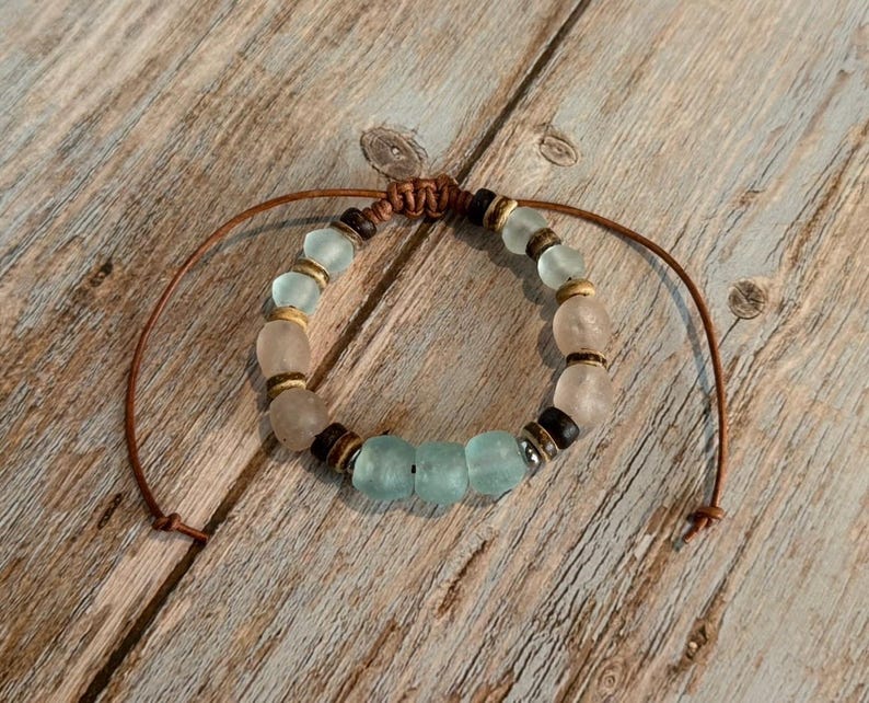 K&ouml;nnte beinhalten: Ein handgefertigtes Armband mit einer Mischung aus hellblauen, klaren und braunen Perlen. Das Armband ist an einer braunen Kordel mit einem Schiebeknotenverschluss befestigt. Die Perlen haben verschiedene Formen und Gr&ouml;&szlig;en und schaffen einen einzigartigen, b&ouml;hmischen Stil.
